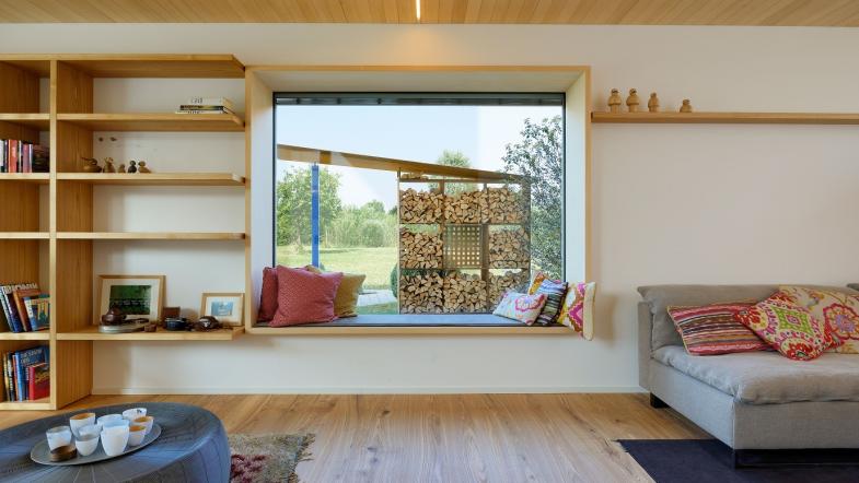 Blick aus Wohnraum durch ein großes Sitzfenster in den Garten auf aufgeschichtetes Holz